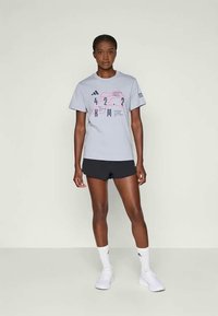 Eine Frau steht vor einem schlichten Hintergrund und trägt ein graues Adidas-T-Shirt mit Grafik, schwarze Shorts, weiße Socken und weiße Turnschuhe.