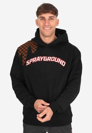 Giovane uomo sorridente, indossa una felpa nera con cappuccio con la scritta “SPRAYGROUND” e motivo a goccia a quadretti marrone sulla spalla, mani giunte davanti.