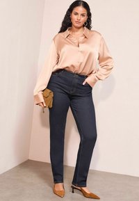 Camicia button-up in satin beige con maniche lunghe, abbinata a jeans blu scuro e décolleté marroni a punta. Piccola borsa marrone con dettaglio a catena.