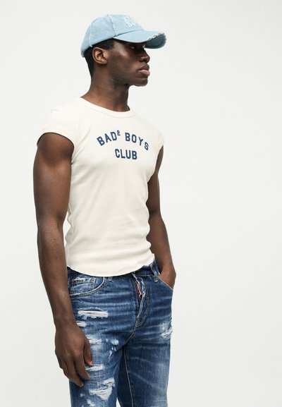 Jeune homme portant une casquette bleu clair, un débardeur blanc avec le texte "BAD² BOYS CLUB", et un jean bleu usé, debout les mains dans les poches.