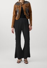 Brązowa skórzana kurtka o krótkim kroju z zapięciem na guziki, czarna koronkowa bluzka pod spodem, szerokie czarne spodnie i czarne sandały na obcasie z paskami.