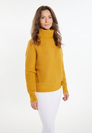 Musztardowa żółta sweter turtleneck z dzianiny o prążkowanej fakturze i długimi rękawami, w połączeniu z białymi dopasowanymi spodniami.