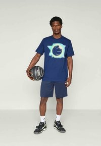 Camiseta azul marino con un diseño gráfico circular, combinada con pantalones cortos azul marino. El modelo sostiene un balón de baloncesto y lleva zapatillas deportivas negras con detalles.