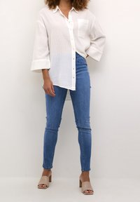 Camisa blanca de textura, de mangas largas, con cierre de botones y bolsillo frontal; jeans ajustados azules; sandalias de tacón beige con punta abierta.