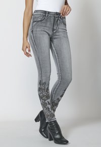 Úzké džíny z vybledlého šedého denimu, s černými vyšívanými květinovými detaily na kotníku. Spárované s černými botami na vysokém podpatku.