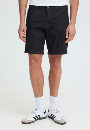 Homme portant un short en jean noir, des chaussettes blanches et des baskets blanches à rayures noires, debout sur un fond blanc uni.