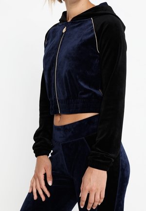 Frau trägt eine marineblaue und schwarze Velours-Cropped-Zip-Hoodie mit passenden hochtaillierten Leggings, steht mit entspannten Händen an den Seiten.