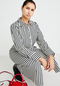 Prúžkovaná dlhá košeľová šaty v čiernej a bielej farbe, s gombíkovým zapínaním vpredu, golierom a opaskom v páse. Doplnkom je malá červená kabelka.