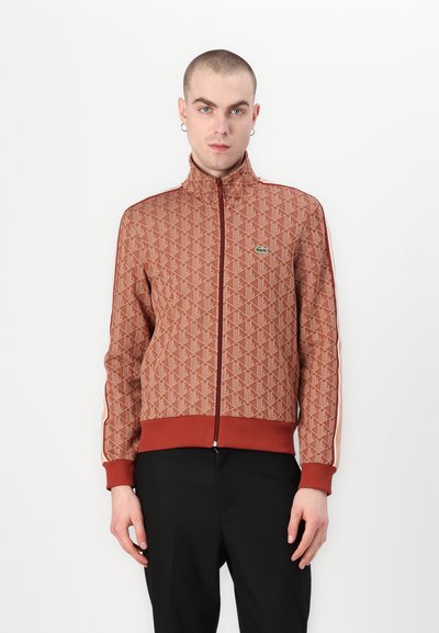 Veste zippée à motif rouge avec un col montant, des poignets et un ourlet élastiques ; présente des bandes blanches contrastantes le long des manches et un logo vert.
