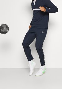 Granatowy dres sportowy z białymi detalami, składający się z bluzy z ćwiartkowym zamkiem i zwężanych spodni. Noszony z białymi i neonowozielonymi korkami do piłki nożnej.