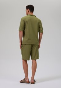 Set de cămașă polo de culoare verde măslin și pantaloni scurți, realizat dintr-un material texturat. Croială lejeră, cu mâneci scurte și guler plat. Asortat cu sandale maronii.