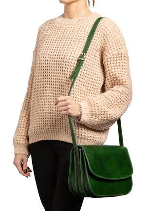 Femme portant un pull en tricot beige et un pantalon noir, tenant un sac bandoulière en cuir vert avec une sangle réglable.