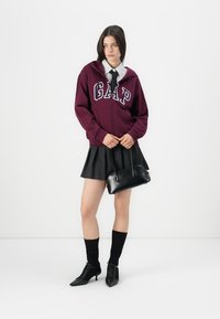Woman wearing a burgundy GAP hoodie over a striped shirt with black tie, black pleated skirt, black socks, heels, holding a black handbag.