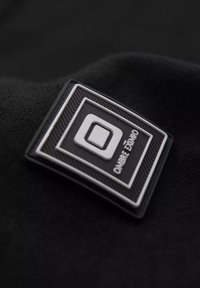 Tejido negro con una etiqueta rectangular en blanco y negro que presenta un logo cuadrado y el texto "OMBRE EXTRIM". Superficie texturizada con acentos en relieve.