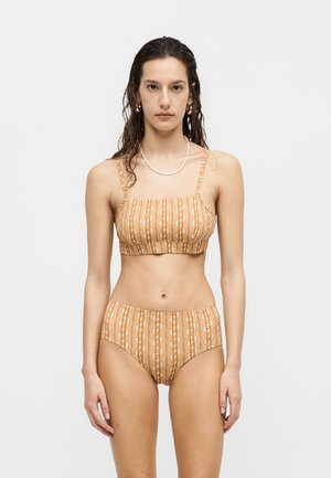 Γυναίκα που στέκεται μπροστά φοράει πορτοκαλί μπικίνι με σχέδιο και σλιπ με ψηλή μέση, διακοσμημένη με χρυσούς κρίκους στα σκουλαρίκια και λευκό περιδέραιο.