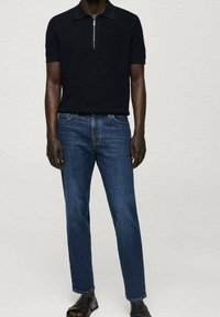 Polo negro de manga corta con collar de cremallera, combinado con jeans azules. Los jeans tienen un corte recto y son ligeramente ajustados en los tobillos. Sandalias negras.