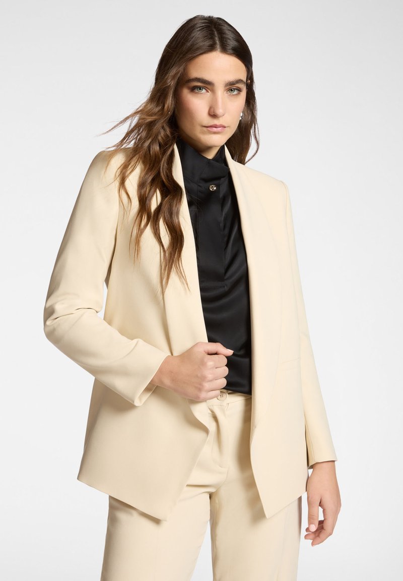 Blazer beige entallado con solapa de muesca, hecho de un material liso. Se lleva sobre una blusa negra de cuello alto, con detalle de botones.