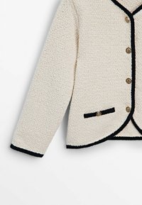 Cardigan à texture crème avec bordure noire, boutons dorés sur le devant et sur une petite poche avant, manches longues et encolure en V.