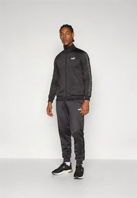 Melns sporta tērps ar rāvējslēdzēju un savienojumu biksēm, uz kurām ir PUMA zīme. Materiāls ir gluds, ar kontrastējošām detaļām uz piedurknēm.