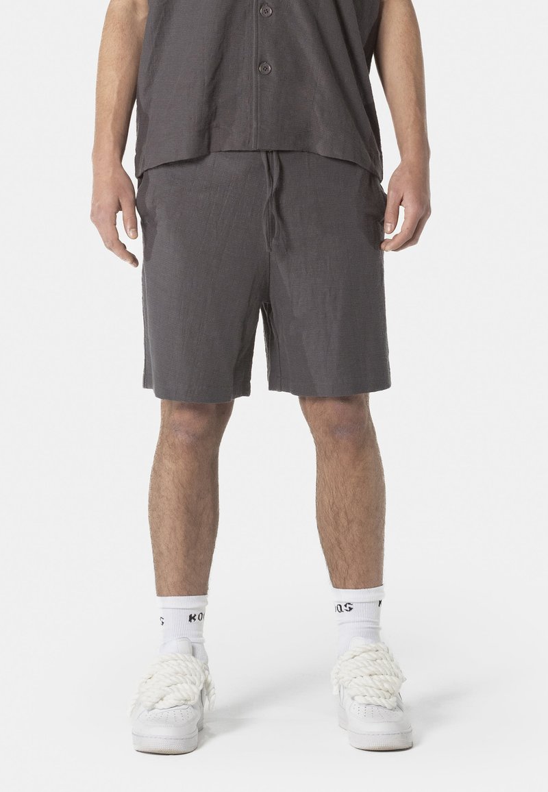 Graue Leinen-Shorts, knielang mit einem Kordelzugbund. Kombiniert mit weißen Turnschuhen und lässigen Socken, dazu ein locker sitzendes Hemd mit Knöpfen.