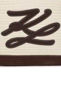 Brūnas ādas logo uz teksturēta krēmkrāsas auduma fona; satur kontrastējošas šuves un vienotu brūnu pamatni.