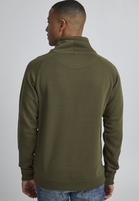Felpa verde oliva con colletto alto, polsini e orlo a costine. Tessuto morbido e testurizzato con dettagli sulle cuciture posteriori. Indossata con jeans blu.