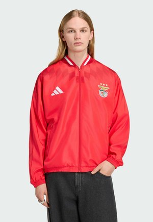 Persona con capelli biondi lunghi indossa una giacca sportiva rossa con zip, logo bianco Adidas e stemma del club calcistico Benfica, abbinata a pantaloni scuri.