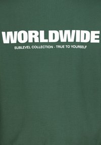 Groene katoenen sweatshirt met vetgedrukte witte tekst: "WORLDWIDE" bovenaan en "SUBLEVEL COLLECTION - TRUE TO YOURSELF" daaronder.