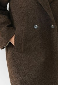 Käsi tummanruskeassa, teksturoidussa villakoissa, jossa näkyy kaksi mustaa nappia ja leveä kaulus.