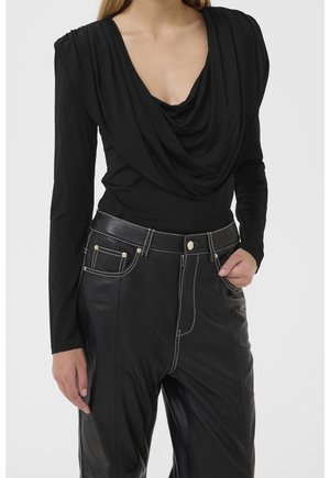 Vrouw draagt een zwarte lange mouw gedrapeerde top en hoge taille zwarte leren broek met witte stiksels en voorzakken.