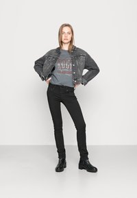 Jovem mulher está de pé com as mãos na cintura, vestindo jeans pretos, botas pretas, uma t-shirt cinza de festival de rock e uma jaqueta de ganga preta curta.