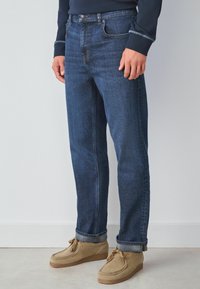 Ciemnoniebieskie dżinsy z denimu o prostym kroju, z pięcioma kieszeniami, zapięciem na guzik i subtelnymi detalami przeszyć. Noszone z beżowymi mokasynami.