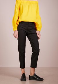 Osoba oblečená v jasně žluté off-shoulder blůze, černých krátkých kalhotách a černých šněrovacích teniskách na neutrálním pozadí.