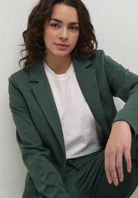 Groene blazer en bijpassende broek over een wit t-shirt. De stof is glad met een gestructureerd ontwerp en een gekartelde revers. Handen rusten op de schoot.