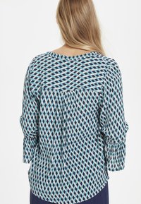 La blusa blu con motivo presenta un design tessuto nei toni del blu navy, blu chiaro e bianco. Ha un collo rotondo e maniche arricciate.