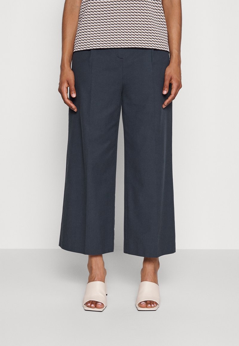 Szerokie, ciemnoniebieskie spodnie wykonane z gładkiego materiału, z dopasowanym krojem i pliskami. Połączone z jasnoróżowymi klapkami z odsłoniętą palcami.