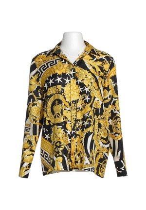 Camisa de manga larga abotonada con patrones florales y geométricos barrocos en negro, blanco y dorado sobre un busto de maniquí blanco.