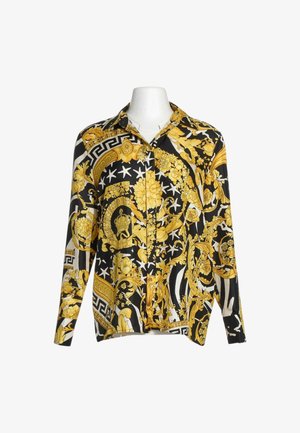 Camisa de manga larga abotonada con patrones florales y geométricos barrocos en negro, blanco y dorado sobre un busto de maniquí blanco.