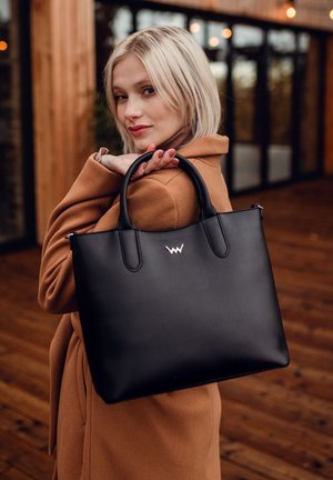 Schwarze Ledertasche mit doppelten Griffen, geprägtem Logo, glatter Textur und strukturiertem Design, getragen über einem beigen Mantel. Holzhintergrund.