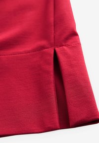 Gros plan sur un tissu de vêtement rouge montrant une couture cousue et une petite fente latérale sur un matériau à texture lisse.