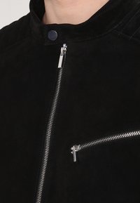 Giacca nera in suede con colletto alto, zip argentate e tasca sul petto. Texture liscia con dettagli di cucitura sottili.