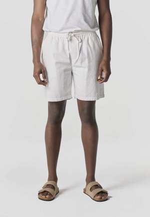 Persona con pantalones cortos blancos con cordón, camiseta blanca y sandalias beige, de pie frente a un fondo claro y sencillo.