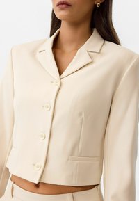 Blazer court beige en tissu lisse, avec un col à revers, une fermeture à cinq boutons et deux poches avant, au design élégant.