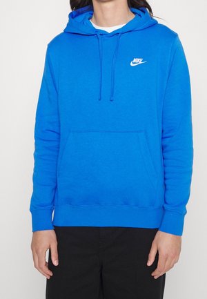 Personne portant un hoodie Nike bleu vif avec une poche avant et des cordons, associé à un pantalon noir, sur un fond blanc uni.
