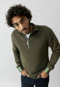 Olive green zip-up sweater with ribbed texture, featuring a stand collar and light green accents on the cuffs. Model poses in a neutral background.
