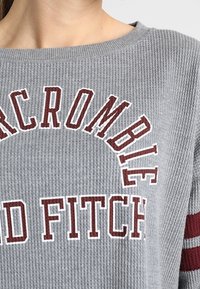 Szary sweter z prążkowanym wzorem, na którym znajduje się biały i bordowy napis "Abercrombie" oraz "Fitch", z bordowymi paskami na rękawach.