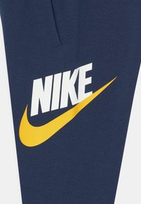 Marinblåa träningsbyxor med vit "NIKE" text och en gul swoosh-logotyp. Tillverkade av strukturerad tyg med sidofickdetalj.