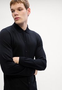 Μαύρη μακρυμάνικη πόλο μπλούζα από πλεκτό ύφασμα, με γιακά, κουμπάκι και εφαρμοσμένα μανίκια. Λείο ύφασμα, ευέλικτος σχεδιασμός.