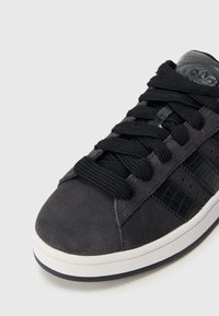 Čierna semišová teniska s textúrovanými čiernymi koženými prvkami, okrúhlymi šnúrkami a bielou gumovou podrážkou. Má perforované detaily na boku.