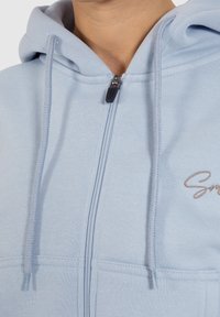Felpa blu chiaro con zip e cappuccio regolabile, caratterizzata da un logo ricamato in un colore discreto, realizzata in un morbido tessuto testurizzato.